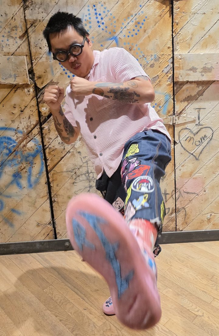 Tri Nguyen throwing a kick at the camera, in glasses and a pink shirt